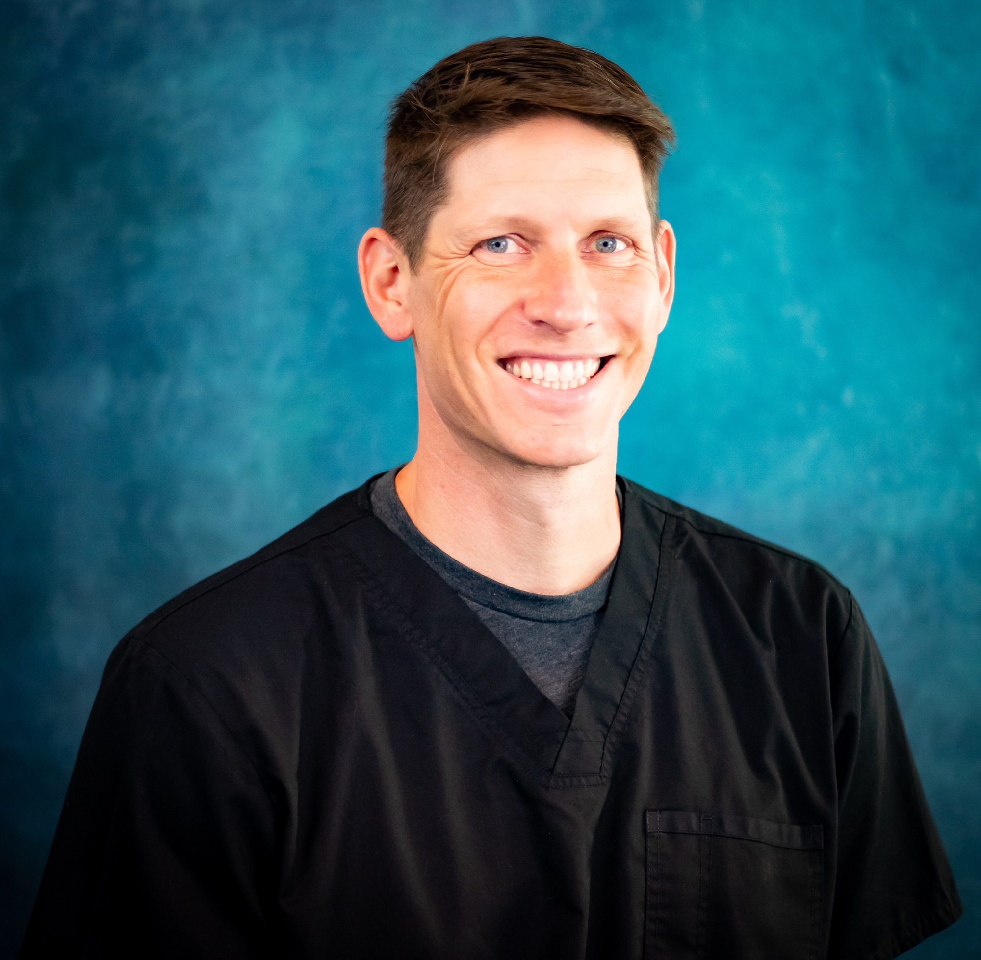 Dr. Jason Rice, orthodontist at Rice Orthodontics, smiling in a black scrub top, against a teal background, representing Jacksonville orthodontics and family orthodontic services.